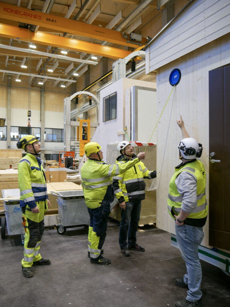 Opiskelijat, opettaja ja työnohjaaja tarkastavat rakennetun testitalon työnjälkeä. Opiskelija mittaa etäisyyttä ilmanvaihtokanavaan.