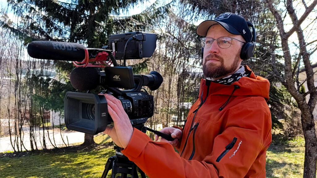 Sivusta otettu kuva jutun päähenkilöstä kuvaamassa videokameralla ulkona. Kuvauskohdetta ei näy.