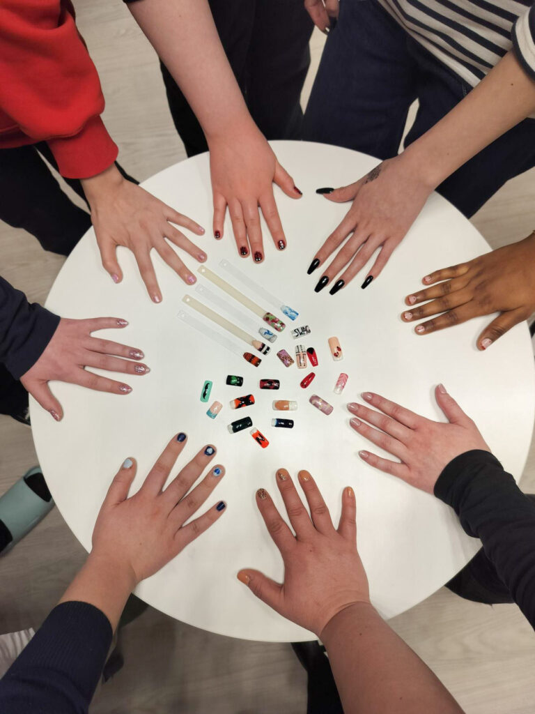 Kahdeksan henkilön käsi on pöytää vasten niin, että kestolakatut kynnet näkyvät. Keskellä on kestolakattuja irtokynsiä.