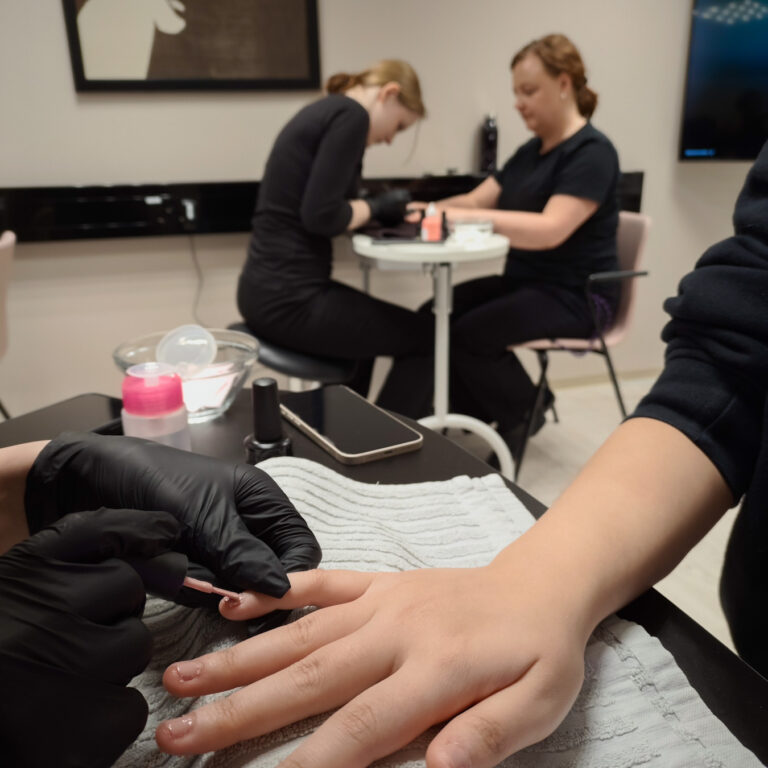 Edessä lakataan kynsiä niin, että vain lakattavan käsi ja lakkaajan kädet näkyvät. Taustalla henkilö tekee toiselle kynsihoitoa.