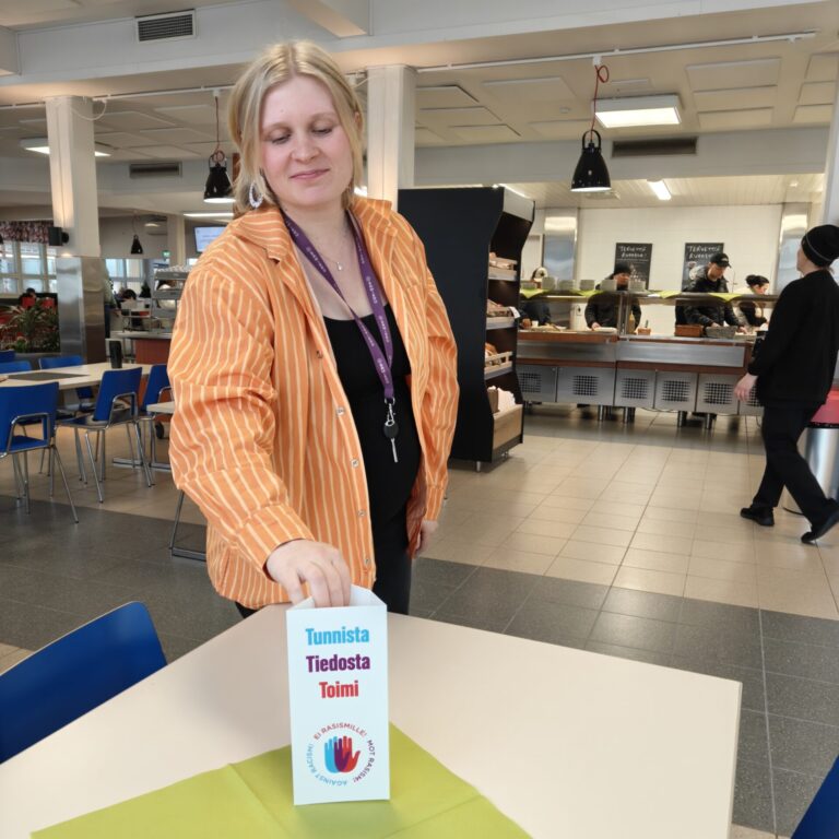 Harjoittelija nostaa ruokalan pöydälle rasisminvastaisen viikon esitettä.