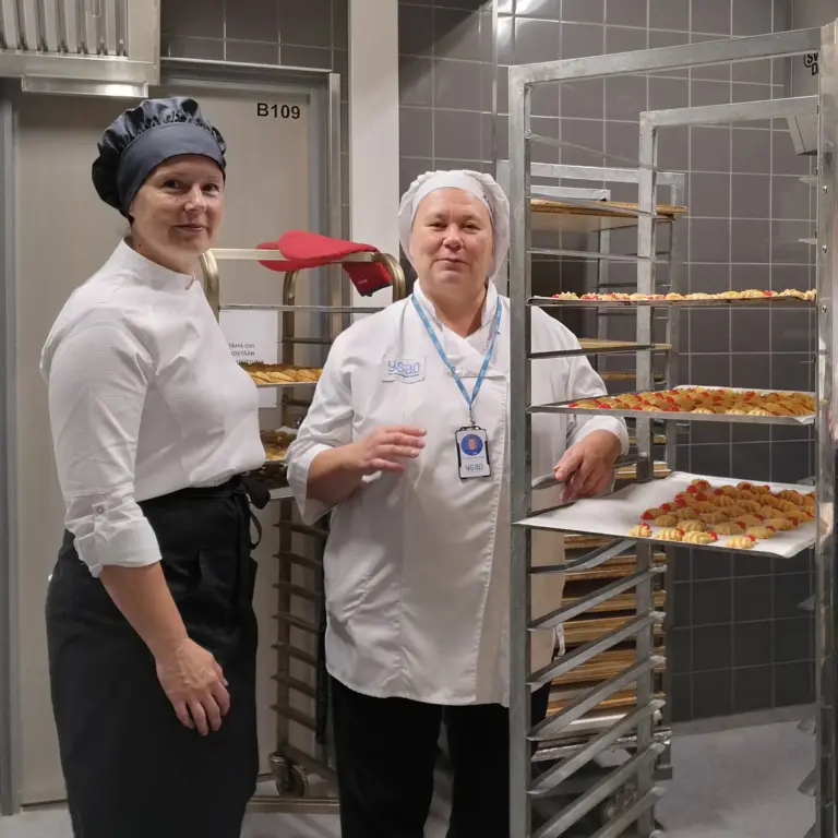 Kaksi leipomoalan opettajaa seisoo paistotelineen vieressä. Telineessä on kaksi pellillistä pikkuleipiä.