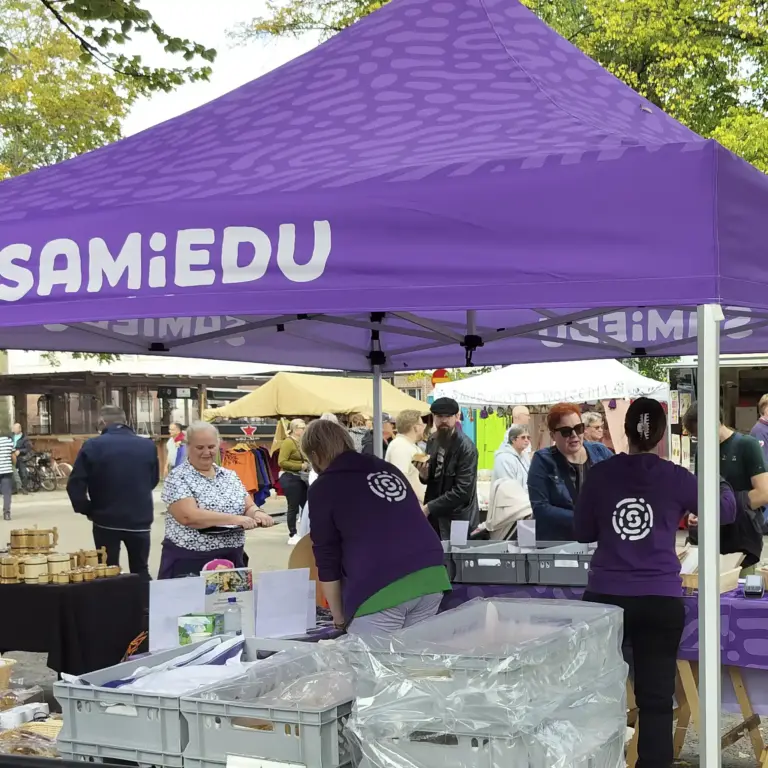 Samiedun violetti teltta toritapahtumassa. Etualalla on leipomotuotteiden laatikoita, teltalla kaksi Samiedu-huppariin pukeutunutta henkilöä asiakaspalvelussa. Taustalla näkyy toriväkeä.