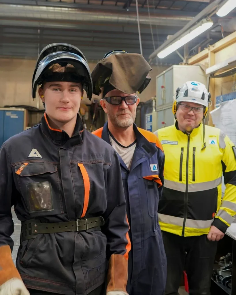 Puolivartalokuvassa on kolme henkilöä, joilla on konepajan työasut ja suojavarusteet.