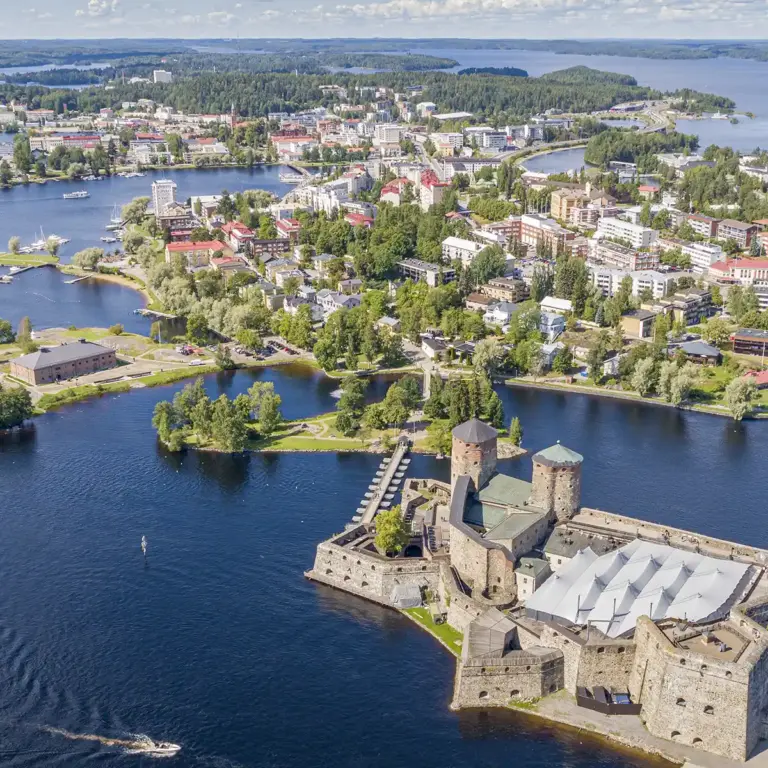 Ilmakuvassa etualalla on osa Olavinlinnaa, järvimaisemaa ja taka-alalla kaupunkia. Kuva on otettu kesällä aurinkoisena päivänä.