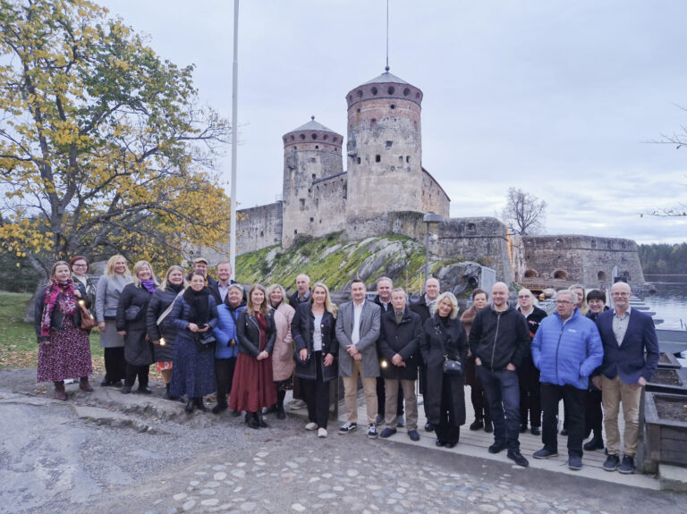 Kuvassa on suuri joukko henkilöitä yhteiskuvassa Olavinlinnan edustalla. Taustalla on pilvinen taivas ja etualalla hiekkatietä.
