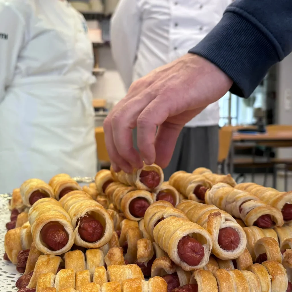 Kuvassa etualalla paistettuja taikinaan käärittyjä nakkeja ja käsi, joka nostaa yhtä kääröä. Taustalla on kahden valkoiseen leipurintakkiin pukeutuneen henkilön hahmot.